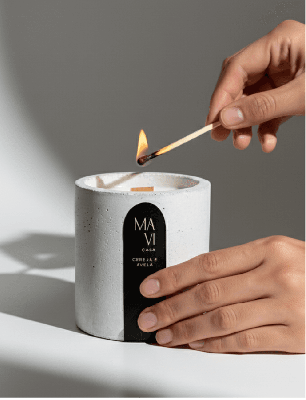 Mãos acendendo com um fósforo o pavio de madeira de uma vela Mavi Casa em pote de concreto branco