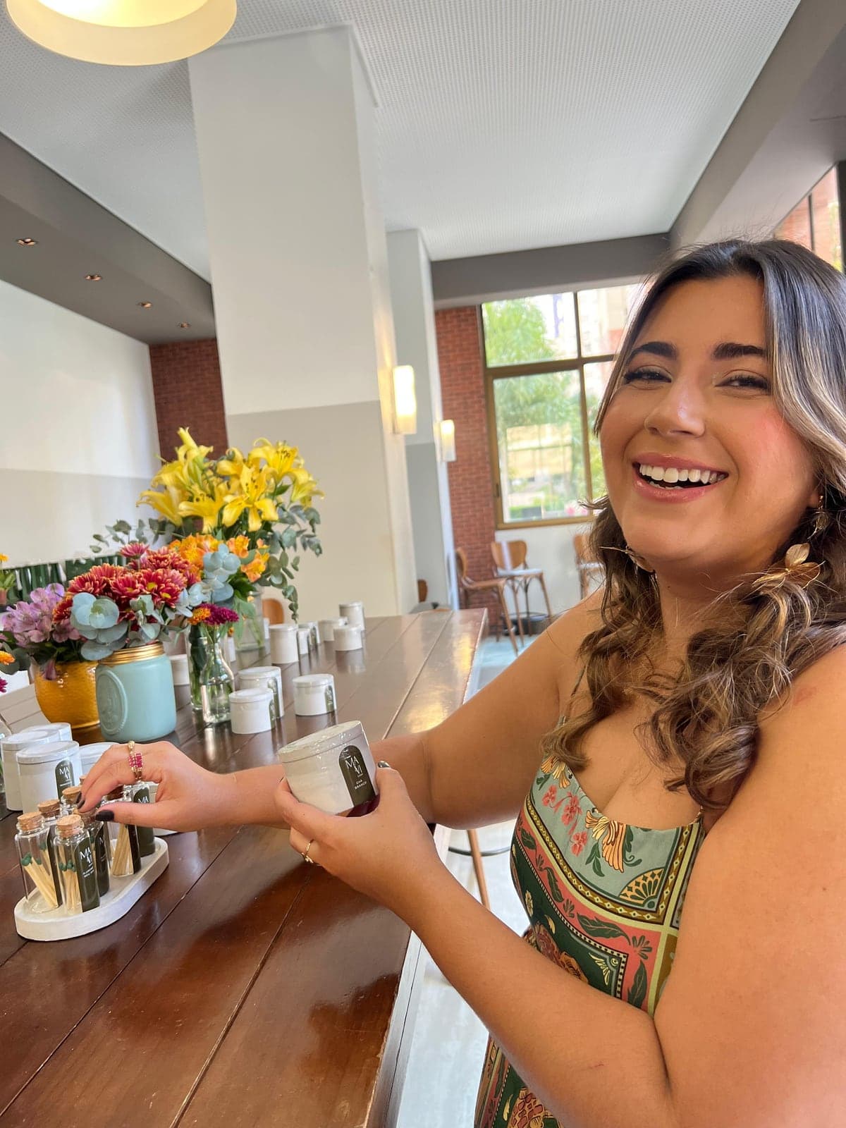 Marina, criadora da Mavi Casa, sorrindo enquanto segura uma vela em pote de concreto sobre uma mesa com arranjos de flores e potinhos do Kit Acenda